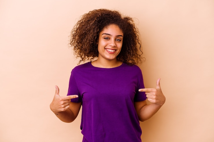 Foto jovem afro-americana isolada em uma parede bege pessoa apontando com a mão para um espaço de cópia de camisa, orgulhosa e confiante