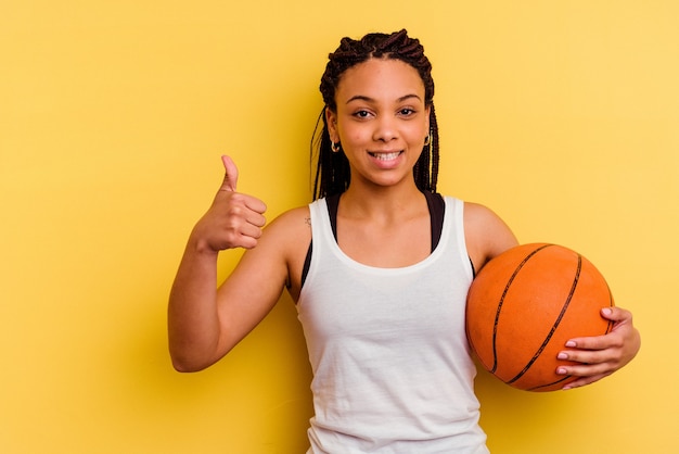 Jovem afro-americana jogando basquete isolado em um fundo amarelo, sorrindo e levantando o polegar