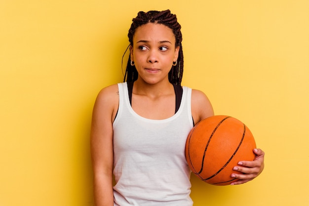 Jovem afro-americana jogando basquete isolado na parede amarela, confusa, duvidosa e insegura