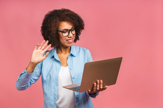 Foto jovem afro-americana negra positiva legal com cabelo encaracolado usando laptop e sorrindo isolada sobre fundo rosa