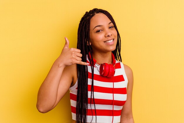 Jovem afro-americana ouvindo música com fones de ouvido isolados na parede amarela, mostrando um gesto de chamada de celular com os dedos