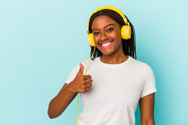 Jovem afro-americana ouvindo música isolada em um fundo azul, sorrindo e levantando o polegar