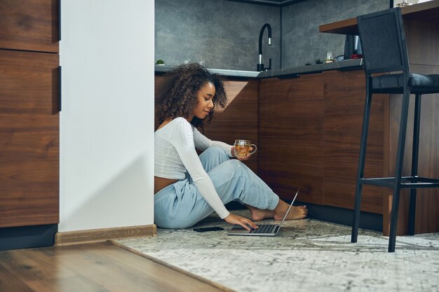 Jovem afro-americana passando um tempo na cozinha enquanto trabalha no laptop e bebe chá