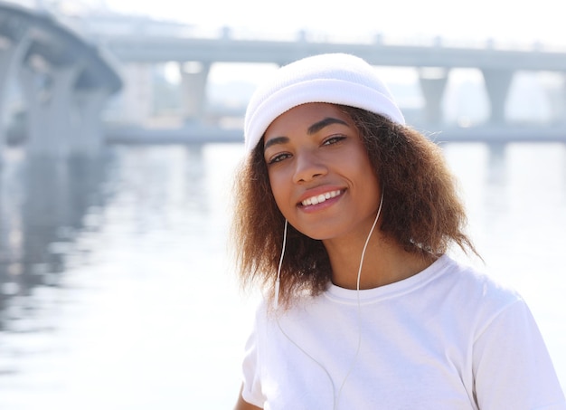 Jovem afro-americana perto do mar e ponte usando telefone celular e fones de ouvido