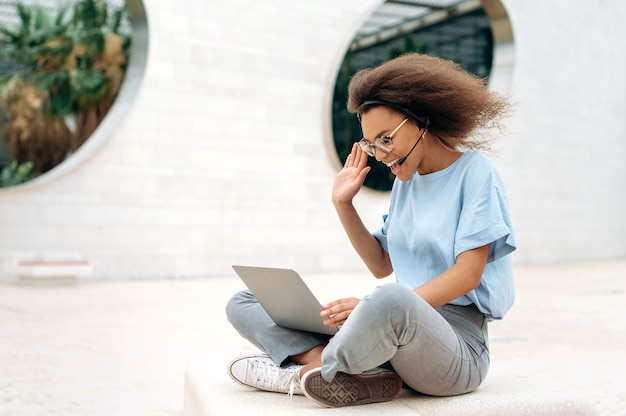 Jovem afro-americana positiva com fone de ouvido realiza consulta on-line distante conversando com colegas ou cliente senta-se ao ar livre usando sorrisos de laptop Conceito de negociação de vídeo on-line