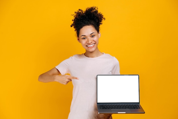 Foto jovem afro-americana satisfeita segurando laptop aberto com tela branca em branco na mão aponta o dedo para ele fica em fundo laranja isolado olhando para o sorriso amigável da câmera conceito de maquete