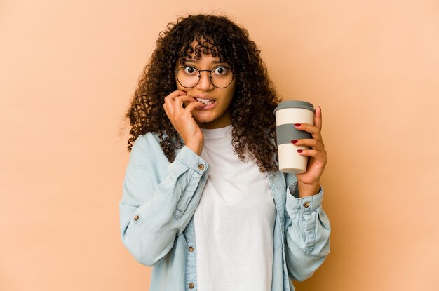 Jovem afro-americana segurando um café para viagem roendo as unhas, nervosa e muito ansiosa.