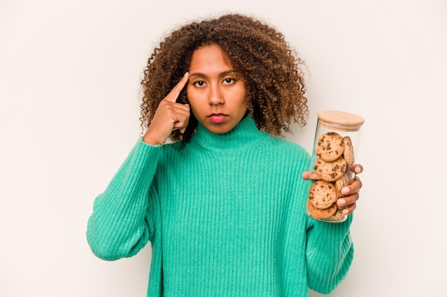 Jovem afro-americana segurando um pote de biscoitos isolado no fundo branco apontando o templo com o dedo pensando focado em uma tarefa