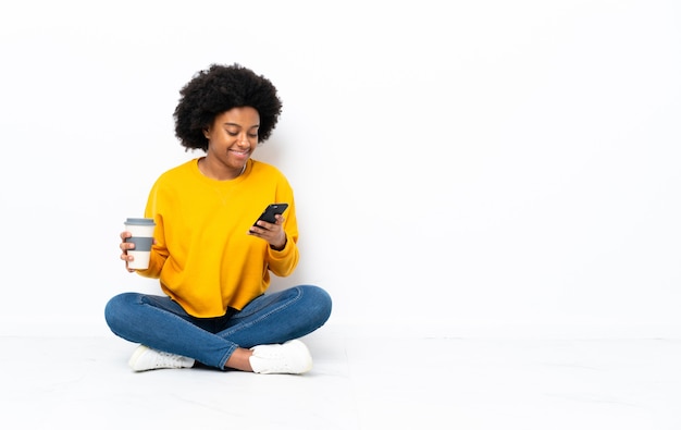 Jovem afro-americana sentada no chão segurando um café para levar e um celular