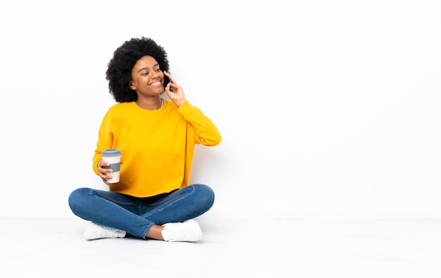 Jovem afro-americana sentada no chão segurando um café para levar e um celular