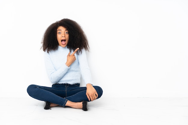 Jovem afro-americana sentada no chão surpresa e apontando para o lado