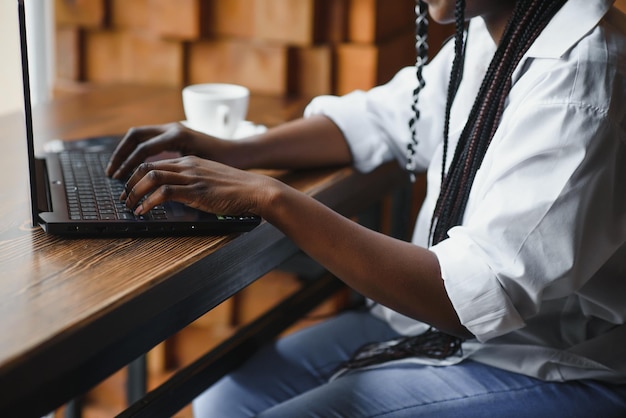 Jovem afro-americana sentada no restaurante e digitando em seu laptop Linda garota trabalhando no computador no café