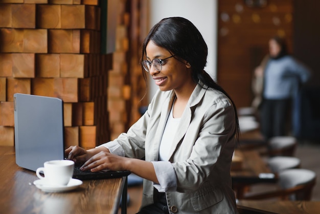 Jovem afro-americana sentada no restaurante e digitando em seu laptop Linda garota trabalhando no computador no café