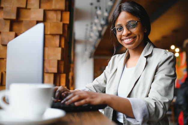 Jovem afro-americana sentada no restaurante e digitando em seu laptop Linda garota trabalhando no computador no café