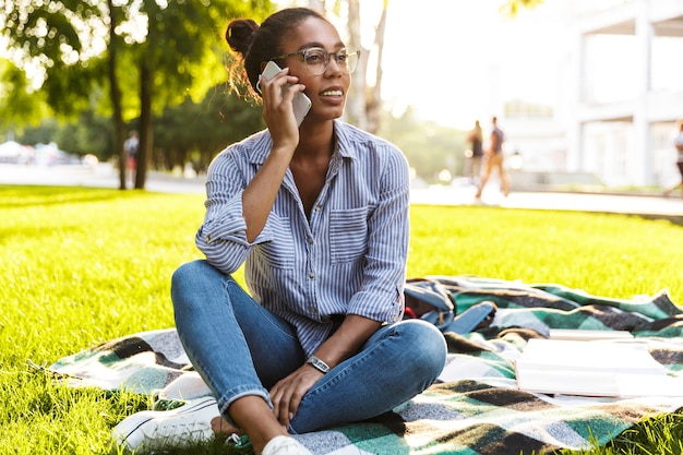 Jovem afro-americana sorrindo e falando no smartphone enquanto está sentada no cobertor no parque