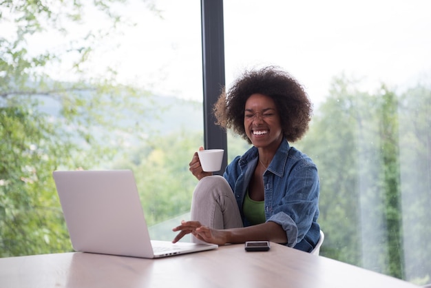 Jovem afro-americana sorrindo sentada perto da janela brilhante enquanto olha para o laptop aberto na mesa e segurando a caneca branca em sua casa de luxo