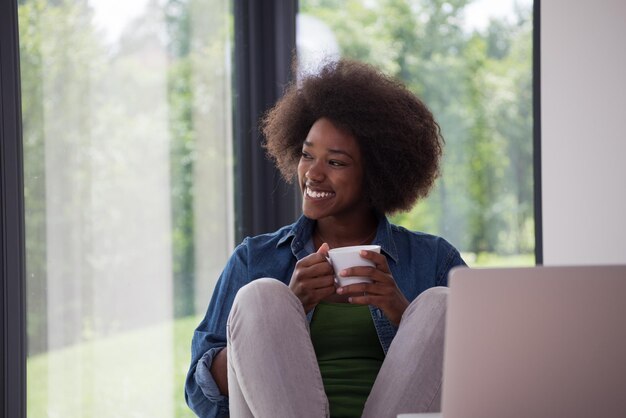 Jovem afro-americana sorrindo sentada perto da janela brilhante enquanto olha para o laptop aberto na mesa e segurando a caneca branca em sua casa de luxo