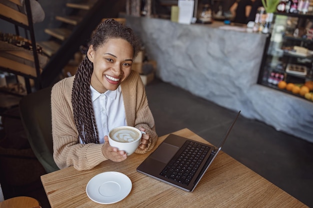 Jovem afro-americana tomando café e usando laptop no café sorrindo lindo sorriso