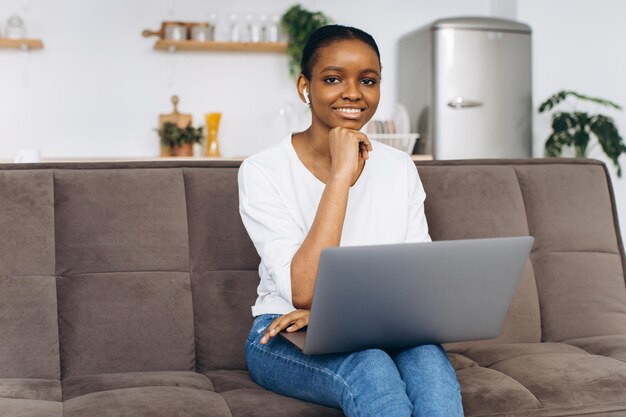 Jovem afro-americana trabalhando no laptop sentado no sofá na cozinha