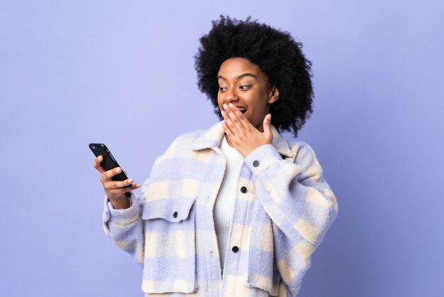 Jovem afro-americana usando um celular isolado em roxo com expressão facial de surpresa e choque