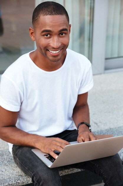 Jovem afro-americano bonito alegre sentado e usando o laptop ao ar livre