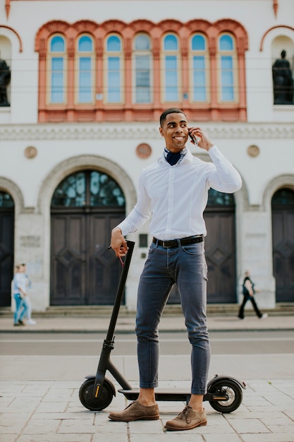 Jovem afro-americano bonito usando um telefone celular em pé com uma scooter elétrica em uma rua