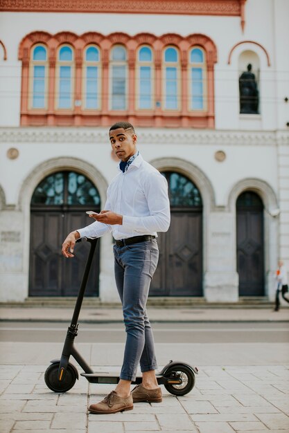 Jovem afro-americano bonito usando um telefone celular em pé com uma scooter elétrica em uma rua