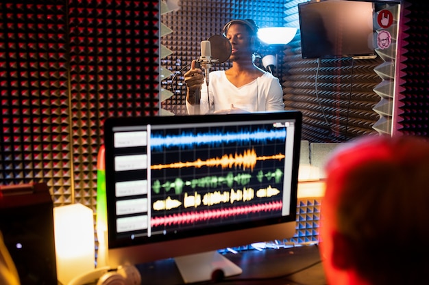 Jovem afro-americano com fones de ouvido cantando no microfone enquanto o produtor grava uma música em frente à tela do computador