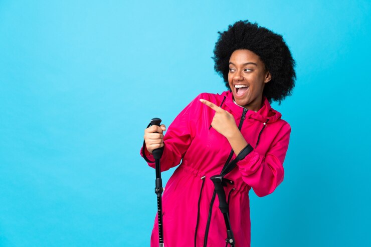 Foto jovem afro-americano com mochila e bastões de trekking isolados em azul apontando o dedo para o lado e apresentando um produto
