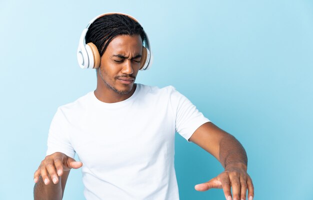 Jovem afro-americano com tranças isoladas em um fundo azul, ouvindo música e dançando