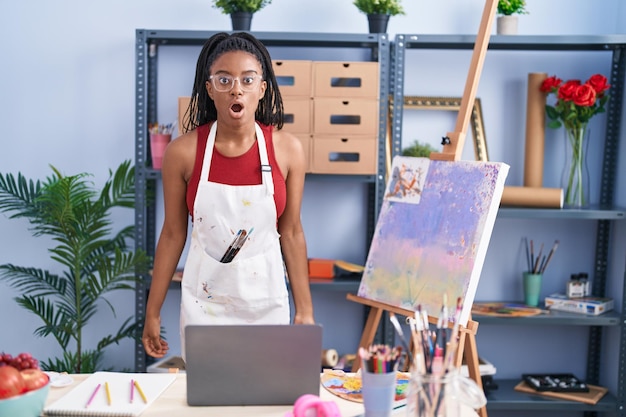 Jovem afro-americano com tranças no estúdio de arte pintando sobre tela assustado e espantado com a boca aberta para surpresa, cara de descrença