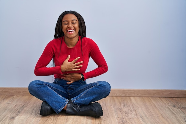 Jovem afro-americano com tranças sentado no chão em casa sorrindo e rindo muito alto porque uma piada maluca com as mãos no corpo.