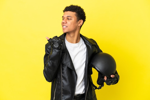 Jovem afro-americano com um capacete de motociclista isolado em um fundo amarelo apontando para o lado para apresentar um produto