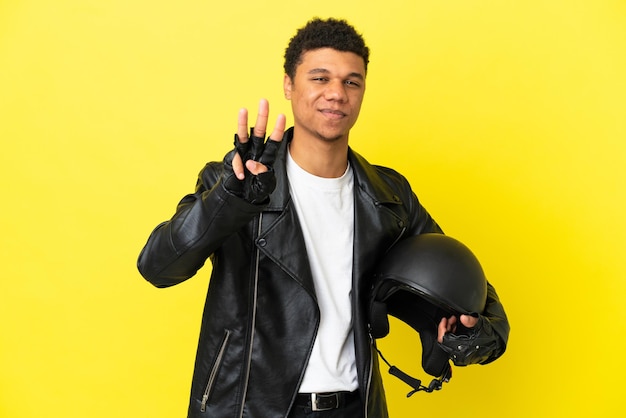 Jovem afro-americano com um capacete de motociclista isolado em um fundo amarelo feliz e contando três com os dedos