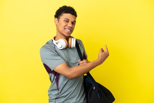 Jovem afro-americano com uma bolsa esportiva isolada em um fundo amarelo apontando para trás