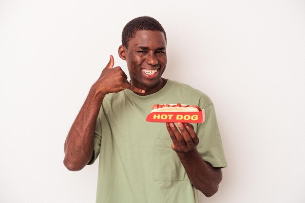 Jovem afro-americano comendo um cachorro-quente isolado no fundo branco, mostrando um gesto de chamada de telefone celular com os dedos