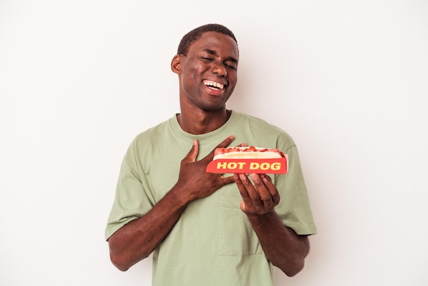 Jovem afro-americano comendo um cachorro-quente isolado no fundo branco ri alto, mantendo a mão no peito.