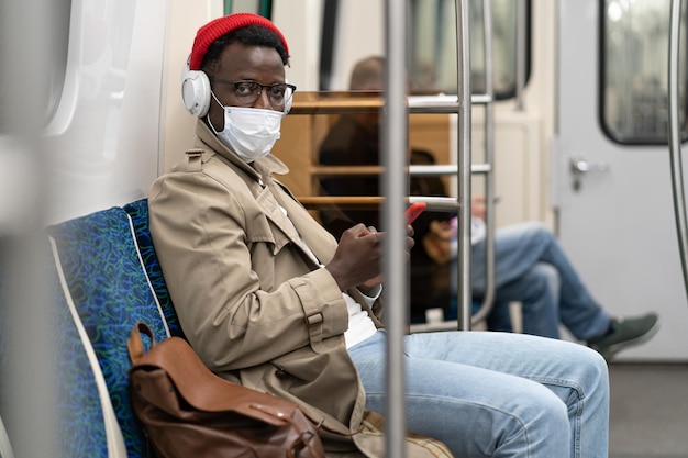 Jovem afro-americano de óculos sentado no metrô usando máscara de rosto ouvindo música
