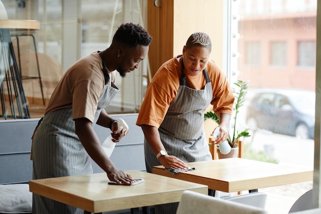 Jovem afro-americano e mulher de avental, trabalhando em um pequeno café, preparando mesas para clientes, limpando-as com spray de detergente e conversando