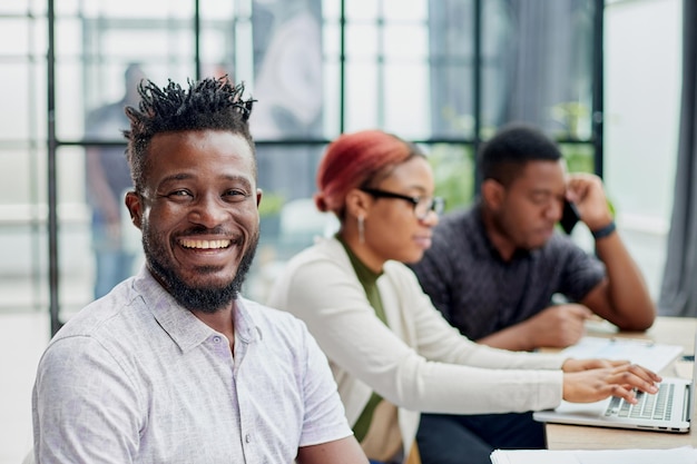 jovem afro-americano em roupas casuais posando para a câmera enquanto está sentado à mesa no contexto de seus colegas