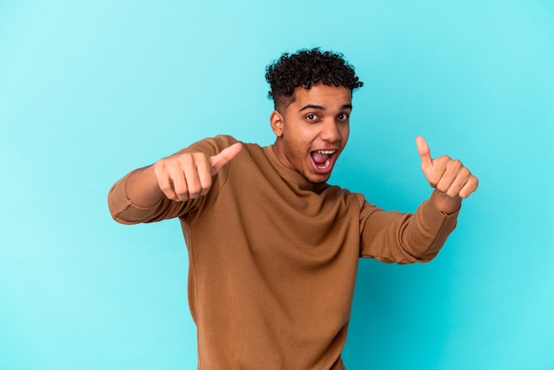 Jovem afro-americano encaracolado isolado em azul, levantando os dois polegares, sorrindo e confiante.