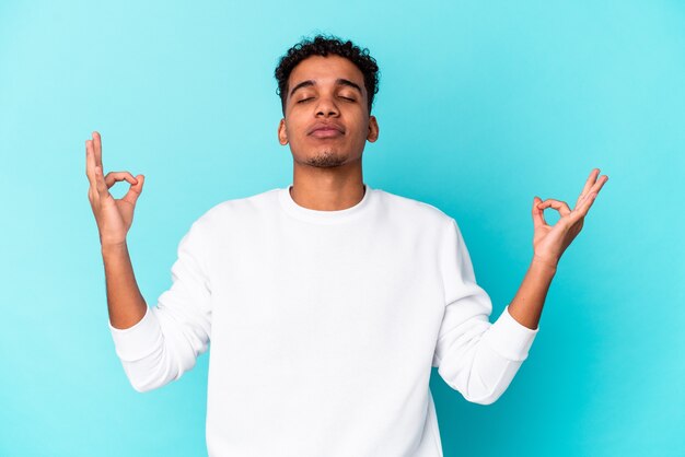 Jovem afro-americano encaracolado isolado em azul relaxa, meditando.