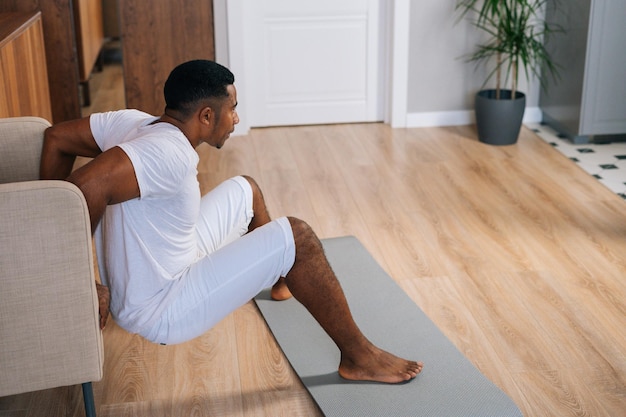 Jovem afro-americano fazendo flexão reversa para os músculos do tríceps usando poltrona no quarto doméstico