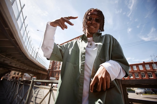 Jovem afro-americano feliz com dreadlocks executando dança hip hop e olhando para a câmera enquanto está de pé contra edifícios modernos