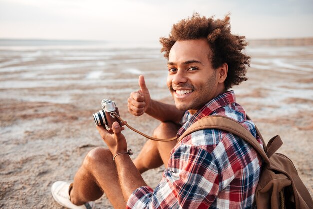 Jovem afro-americano feliz com mochila tirando fotos e mostrando os polegares para cima na praia