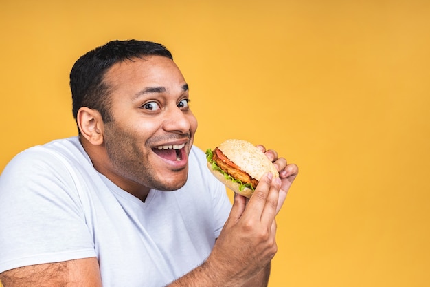 Jovem afro-americano indiano negro comendo hambúrguer isolado sobre fundo amarelo. Conceito de dieta.