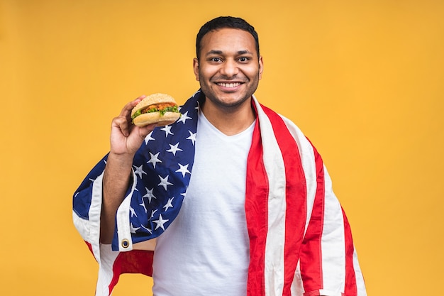 Jovem afro-americano indiano negro comendo hambúrguer isolado sobre um fundo amarelo com a bandeira americana. Conceito de dieta.