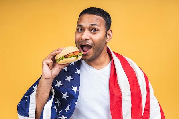 Jovem afro-americano indiano negro comendo hambúrguer isolado sobre um fundo amarelo com a bandeira americana. Conceito de dieta.