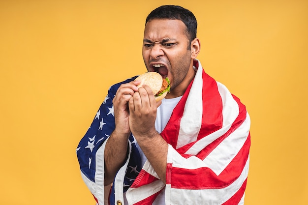 Jovem afro-americano indiano negro comendo hambúrguer isolado sobre um fundo amarelo com a bandeira americana. Conceito de dieta.