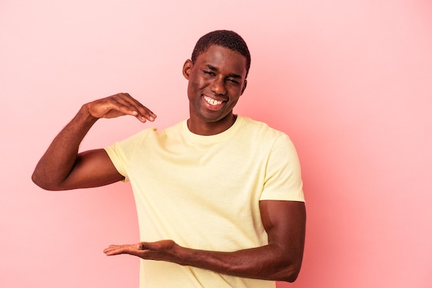 Jovem afro-americano isolado em fundo rosa segurando algo pequeno com os dedos sorrindo e confiante